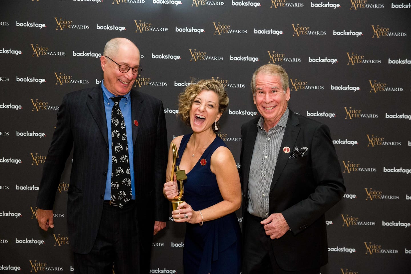 Three guests laughing together on the Voice Arts Awards step-and-repeat, the woman in the middle holding a gold trophy