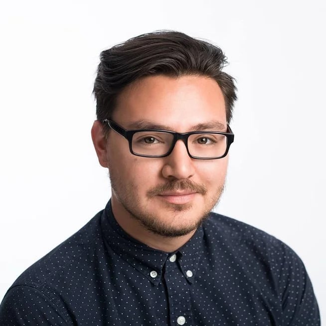 Portrait of a young man with dark hair, wearing glasses and a navy blue polka-dot shirt against a white background